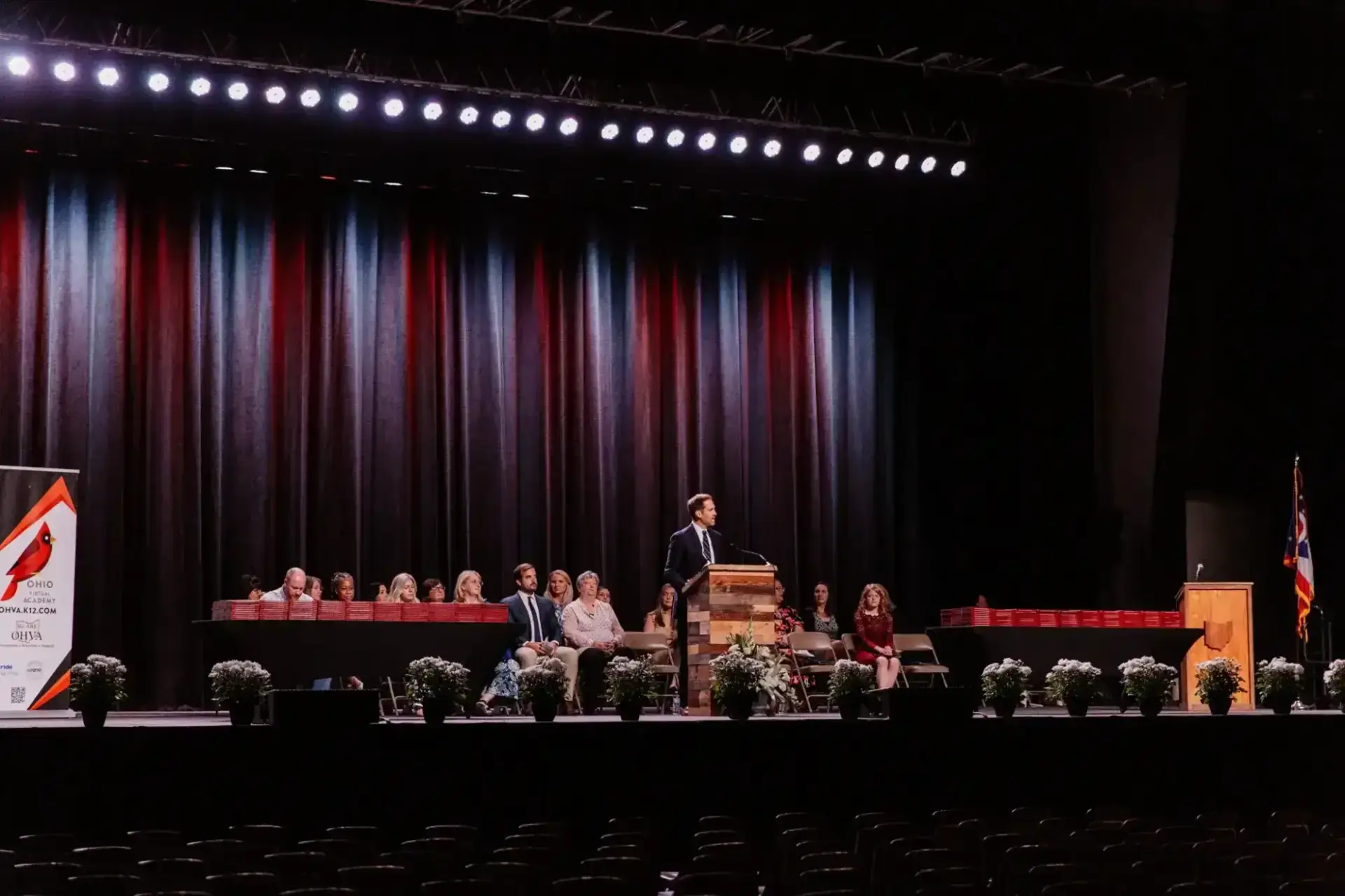 Nin ayaa soo istaagay goob madal ah oo ku taal masrax weyn oo leh daah, isaga oo la hadlaya dhegaystayaashii. Dadka fadhiya iyo dhirta dheriga leh ayaa soo taagan masraxa. Jawigu waa mid rasmi ah.